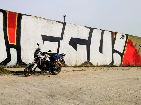 A white adventure motorcycle parked in front of a large graffiti-covered concrete wall with bold black, red, and yellow shapes, on a dusty urban roadside.