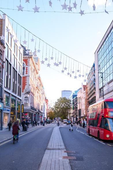 A bustling city street adorned with star-shaped decorations overhead. The scene includes a mix of pedestrians, cyclists, and vehicles, with notable buildings and storefronts lining the street. A red double-decker bus is prominently visible on the right side.