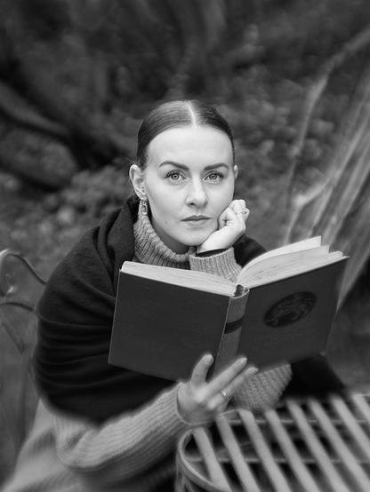 A woman sits outdoors on a chair next to a round table with a grid pattern. She wears a turtleneck sweater and a dark shawl or wrap. She holds an open book in her left hand and rests her chin on her right hand. Blurred trees and plants are visible in the background.