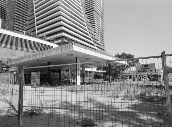 A fenced off, derelict, and decaying gas station occupies space next to shiny, new condominium towers with swooping lines. To the right, a low structure, which was likely the car wash, has block, graffiti letters that declare “BLAME”.