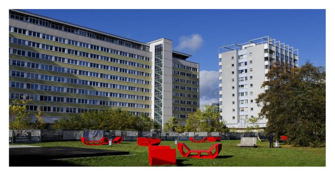 Umrahmt von hohen Plattenbauten eine grüne Wiese, auf der rote Sitzgelegenheiten von der Sonne knallig in Szene gesetzt werden