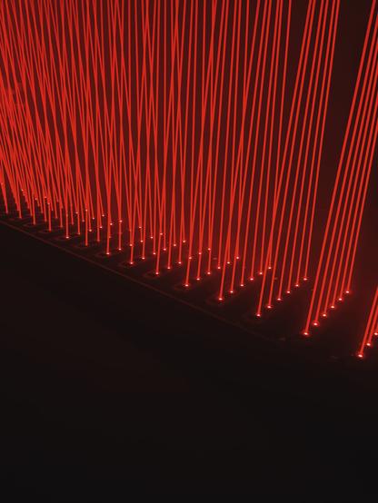 The image shows a dense arrangement of thin, straight red light fibers or laser beams positioned vertically next to each other. These light lines create the impression of a thick, vertical curtain or veil. The light source is located at the bottom, where small dots are visible, emitting the light upward. The surroundings are completely dark, which further emphasizes the intense red color and the striking light effect. The overall impression is futuristic and highly impactful, evoking a modern art installation or a light show. The space appears deep and three-dimensional.