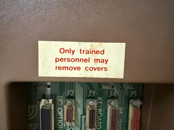 Image shows a close-up of a warning label affixed to a brown surface, advising that "Only trained personnel may remove covers." Below the label, there are visible electronic components, including several connectors and circuit traces.