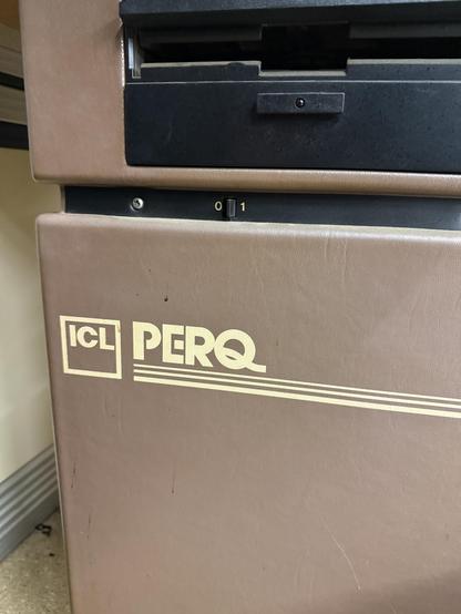 Image shows a close-up of an old computer system labeled "ICL PERQ." The surface is textured and brown, featuring a black section with a small slot. There are also dials or indicators marked with the numbers 0 and 1.