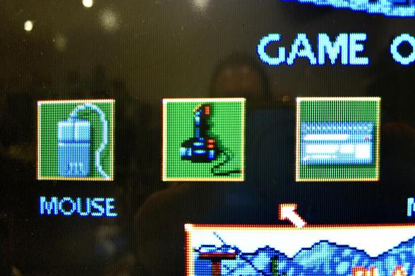A close-up of a CRT TV showing the menu screen for the Amiga game Midwinter, focussing on a row of three icons for the control options. First is a Mouse icon showing an image of a grey mouse with an Atari logo on it, then a Joystick icon showing an image of a black joystick, and finally a Keyboard icon showing an image of a grey computer looking somewhat like an Atari ST.