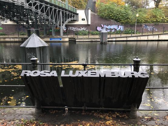 Ein Kunstwerk neben der Brücke. Ein Eisenblock aus der oben den Namen Rosa Luxemburg zeigt.