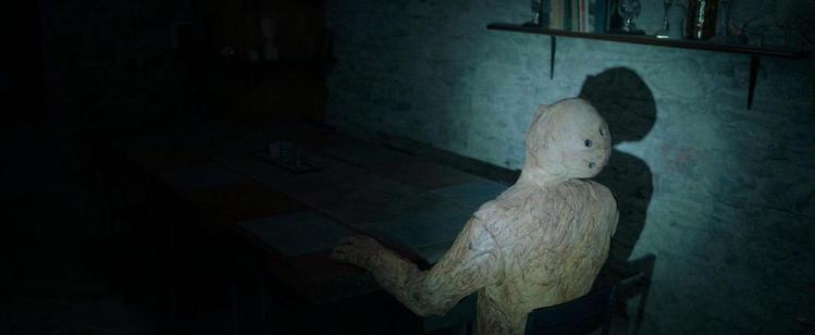 A wooden puppets sits in the dark at a dinner table inside an old stone house. A flashlight shines on it.