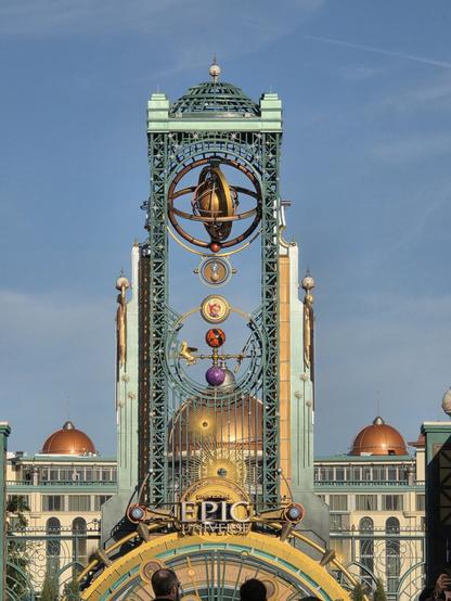 A photograph of the entrance gateway to the Epic Universe theme park at Universal Orlando Resort in Florida. It is a tower, called the Chronos Tower, that has several elaborate designs on it, symbolizing various parts of the park. In the center there is a large, spinning gyroscope with the Time-Turner hourglass from Harry Potter directly underneath it. Under that is a circular emblem depicting Mario, and then further below there are two spheres, I believe representing How to Train Your Dragon and Dark Universe. In between and off to the sides of those spheres there are depictions of a pegasus and comets, representing the Constellation Carousel and Stardust Racers attractions in Celestial Park. Below these designs is a sign that says "UNIVERSAL EPIC UNIVERSE". Behind this tower the Universal Helios Grand Hotel can be seen taking up the bottom quarter of background.
