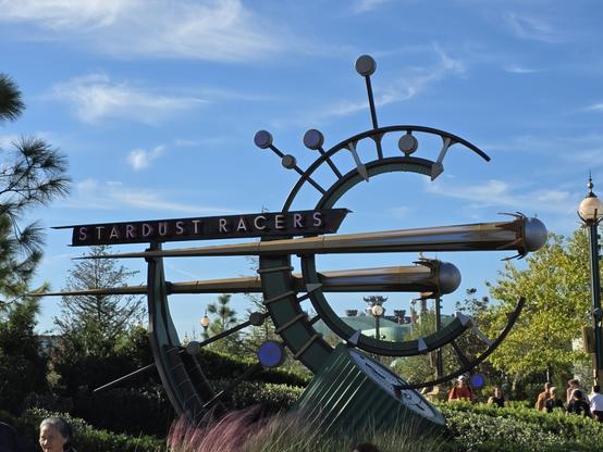 A photograph taken at the Epic Universe theme park at Universal Orlando Resort. It is of a sign for the Stardust Racers attraction in Celestial Park. It has designs depicting shooting stars and a sextant.