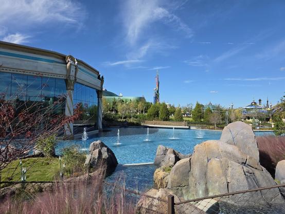 A photograph taken at the Epic Universe theme park at Universal Orlando Resort. It is a shot of a body of water in Celestial Park with several fountains in it past a small waterway flowing into it. On the left the Atlantic restaurant, and on the right is the Constellation Carousel, with the spire of the Super Nintendo Land portal visible in the center.