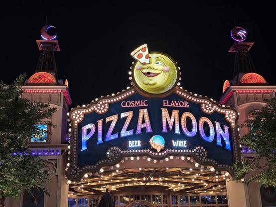 A photograph taken at the Epic Universe theme park at Universal Orlando Resort. It shows the front exterior of the Pizza Moon restaurant located in Celestial Park, taken at night. The sign says "COSMIC FLAVOR - PIZZA MOON - BEER - WINE" and has a moon with a face, which has a slice of pepperoni pizza lodged in its right eye (it seems fine though, it's smiling). The building is supposed to resemble a 1900s Victorian theater and has many lights and two towers on each side, topped with a moon and a shooting star design on the left and right respectively.