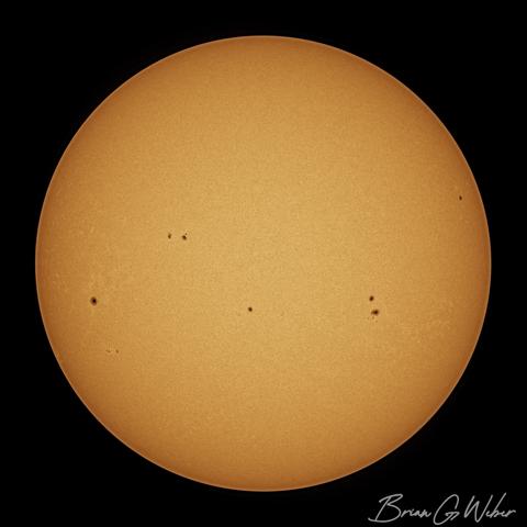 A full disk solar image captured with a monochrome camera and colored orange. The surface is dotted with numerous sunspot clusters all across the visible area.