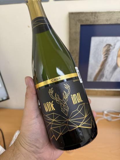 A hand holding a bottle of cava with a black and gold label featuring a geometric deer design and the text "Wine imal." The background shows a wooden surface and some blurred artwork.