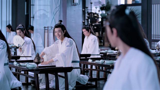 In a classroom in the Cloud Recesses, a mischievous teenage Wei Wuxian grins at a blurred Lan Wangji who has turned to look at him in reprimand. Nie Huaisang is seated to Wei Wuxian's right, his lower face hidden behind a fan. There are a few other unnamed students visible around the room.