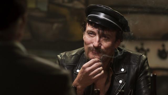 Ein Mann (Oliver Masucci als R.W.Fassbinder) trägt eine schwarze Lederjacke mit silbernen Reißverschlüssen und Knöpfen. Der Mann hält eine Hand mit einer Zigarette vor den Mund. Im Hintergrund sind unscharfe Gegenstände und eine weitere Person zu erkennen. (Bild: © Bavaria Filmproduktion)