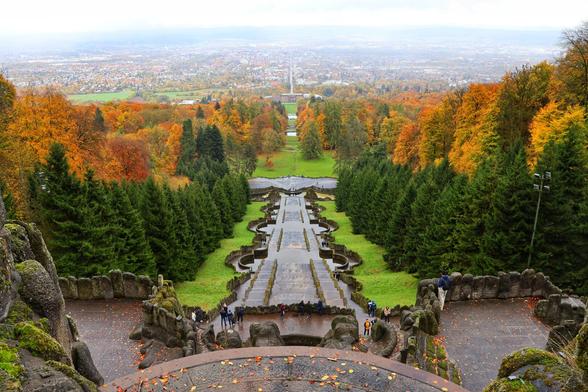 This image presents a sweeping view of Bergpark Wilhelmshöhe, captured from the top of the hill just below the Herkules monument. The scene is dominated by the park’s iconic steep cascades and grand staircases, which descend symmetrically towards the Wilhelmshöhe Palace in the distance. The cascades are framed by dense, autumnal trees, their leaves displaying a vibrant mix of greens, oranges, and reds.

The foreground features a stone terrace with scattered fallen leaves, adding to the seasonal ambiance. A few visitors can be seen exploring the cascades, providing a sense of scale and tranquillity. Beyond the park, the landscape extends into an urban expanse, with buildings and fields stretching towards the horizon under a soft, overcast sky. The image captures the majestic blend of nature, architecture, and history that defines Bergpark Wilhelmshöhe.