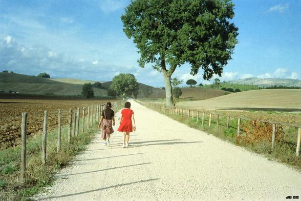 Farbfoto mit zwei Frauen in Bildmitte, die auf einem Schotterweg spazieren. Links und rechts vom Weg ein halbhoher Zaun mit Holzpfosten, weiter entfernt Laubbäume und Hügel. Blauer Himmel mit verstreuten weißen Wolken. Olympus 35LC auf Kodak Gold.
