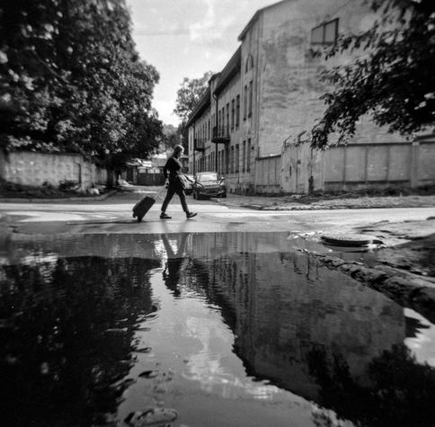 This black-and-white photograph, taken in Lviv with a Holga camera, captures a solitary person crossing a quiet street while pulling a suitcase. The scene unfolds between old, weathered buildings, with trees framing the composition. A large puddle in the foreground reflects both the passerby and the surrounding architecture, creating a striking sense of symmetry and depth. The characteristic soft focus and vignetting of the Holga add a dreamlike, nostalgic quality to the image, evoking a feeling of fleeting journeys and quiet urban moments.