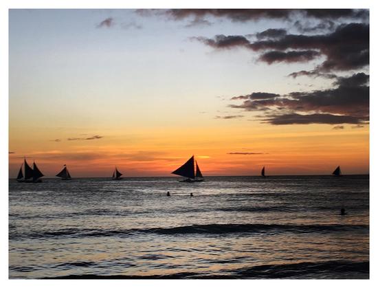 Photo of a sunset over a calm sea. Silhouetted sailboats glide along the horizon. Two distant swimmers poke their heads above the water at the center of the image. A mass of dark clouds pushes in from right frame, but the sky is mostly clear, in a gradient from pale blue to orange-gold.
