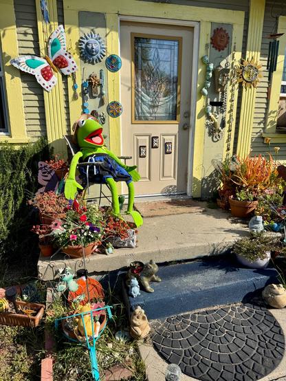 An older house is painted chartreuse yellow and is adorned with a lot of ornamental art consisting of suns, butterflies, and frogs everywhere! On the front porch is a full size chair with a huge kermit the Frog sitting there to great people. Kermit is wearing blue shorts. The front door has a leaded glass window in it and decorative address tiles. There are also potted plants on the porch and front walkway/steps. San Luis Obispo, California.