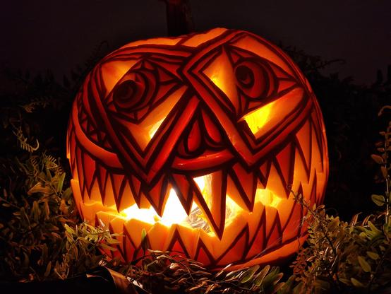 Ornately carved pumpkin. One eye is a square with a triangle iris then round pupil in the middle. The other is a triangle with a square iris and round pupil. The brow line is alternating triangles. It has sharp teeth in a big wide grin, each of the individual teeth have also have triangles carved out, leaving only an outline.