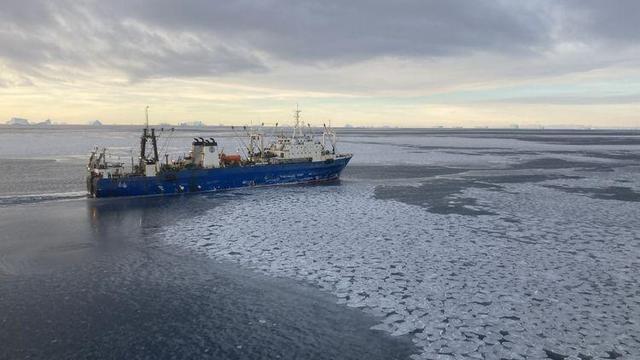 Ein blaues Forschungsschiff mit Kranaufbauten im zugefrorenen Meer, auf der Wasseroberfläche liegt Schnee. Der Himmel ist leicht bewölkt, das Wetter ruhig.