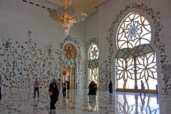 A broad view of a large room, all white except for the flowering vine motif on the floor and on the walls. On the walls, the vines are made of tile and applied to the walls. The windows are massive with a lower section that is rectangular-shaped and the top is like a dome. The windows have the same flower motif. There is an ornate chandelier hanging from the ceiling and a few people are walking around the room. The men are in street clothes/business suits. The women are in black robes.