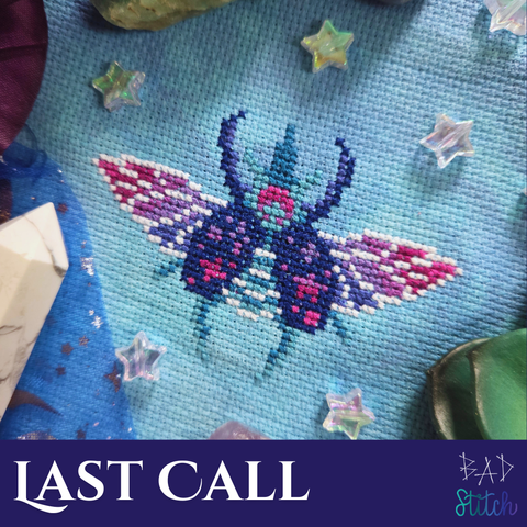 A close up of a cross stitch pattern on hazy blue fabric, depicting a beetle, with wings extended.  There are small starry motifs on the beetle's carapace and a soothing, pink and purple gradient on the wings.  The project is staged with star beads, succulents, and a white tower crystal.  Last Call. BAD Stitch.