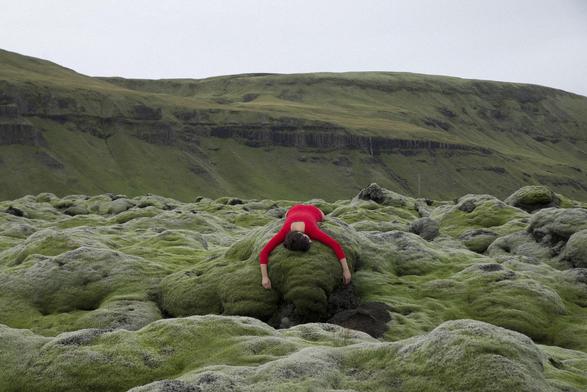 au pied d'une falaise une femme en body rouge couchée à plat ventre sur les rochers couverts d'algues vertes