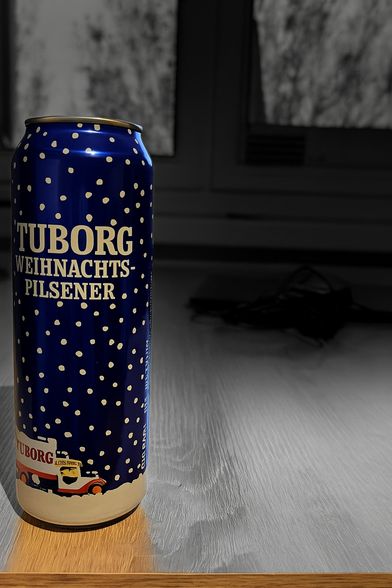 Eine blau-weiße Dose Tuborg Weihnachts-Pilsener(auf dem blauen Grund sind fallende Schneeflocken zu sehen) steht auf einem Holztisch im Vordergrund. Die Dose ist farbig hervorgehoben, während der Hintergrund in Schwarz-Weiß und unscharf dargestellt ist. Im Hintergrund erkennt man ein Fenster und einige Gegenstände auf dem Tisch.