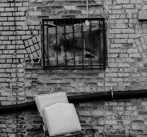 A black and white photo of a very low to the ground barred window in a brick wall. There is corrugated plastic conduit running along the wall underneath and seat cushions stacked on a barely visible chair.