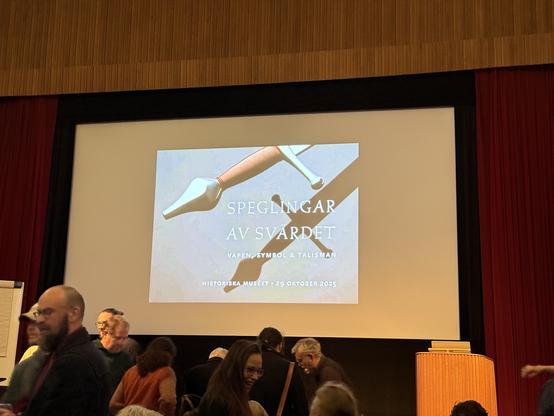 Photo of a screen behind a stage. There is an image of a sword hilt on the screen, along with the words “Speglingar av svärdet — Vapen, symbol & talisman”.