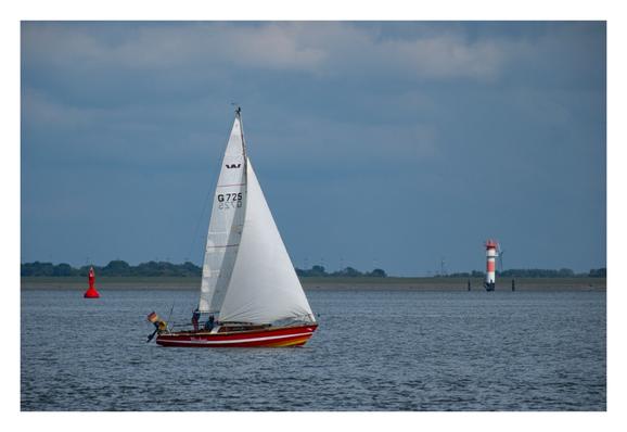 Foto im Querformat. Blick über die Nordsee auf die Küste. Ein rot-weiß gestreiftes Segelboot fährt von links nach rechts, darauf zwei Menschen. Das Boot heißt „Puschen“. Links dahinter schwimmt eine rote Boje im Wasser. Rechts an der Küste ist ein rot-weiß gestreifter Turm. An der Küste an sich sind Wiesen und Wälder. Der Himmel ist bewölkt.