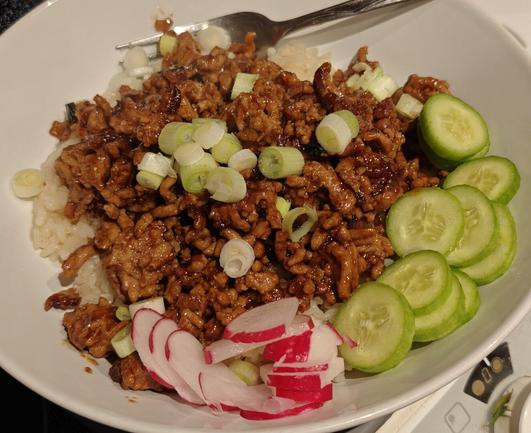 Cooked minced pork in a Thai sweet chilli and basil sauce. Served in a white bowl with rice, and sliced spring onions, radishes, and small cucumbers.