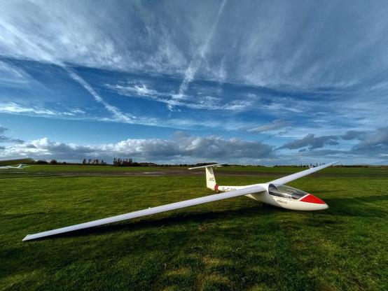 A single seat glider aircraft sits on a grassy field with blue skies and white clouds behind it. The golden light of autumn floods the scene