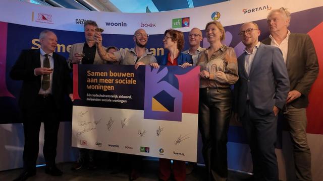 De Vries en Voortman heffen het glas met woon corporatie bestuurders. Ze houden een grote cheque met hun handtekeningen eronder vast