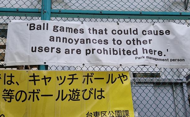 A sign at an outdoor sportcourt that says: ball games that could cause annoyances to other users are prohibited here. (Park management person) Below a sign with the Japanese translation.