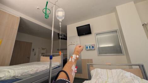 Eine hochgereckte Hand in mit zwei Zugängen für Medikamente im Arm in einem Krankenhauszimmer. An einem Ständer hängt ein Tropf mit einer klaren Flüssigkeit.