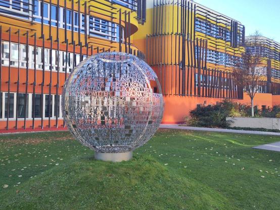 Eine Metallkugel auf einem Grashügel, vor dem bunten Verwaltungsgebäude der Wirtschaftsuniversität Wien.