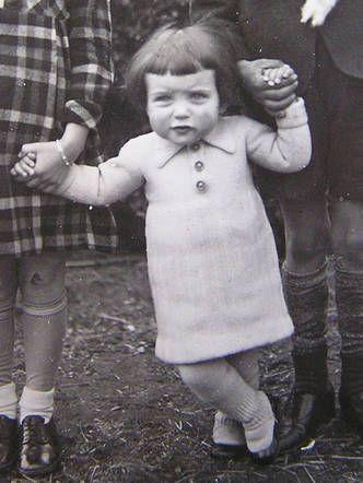 A baby girl standing in her dress - she holds two people by their hands.