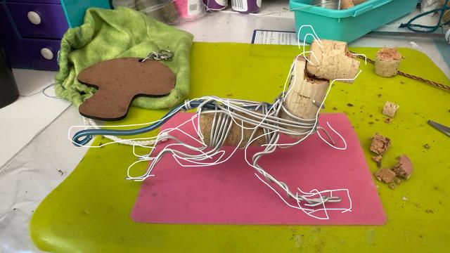 messy craft station with a wire armature in the middle. the general shape of an animal is supported with drawn lines. cork is being used as filler and to make the piece lighter