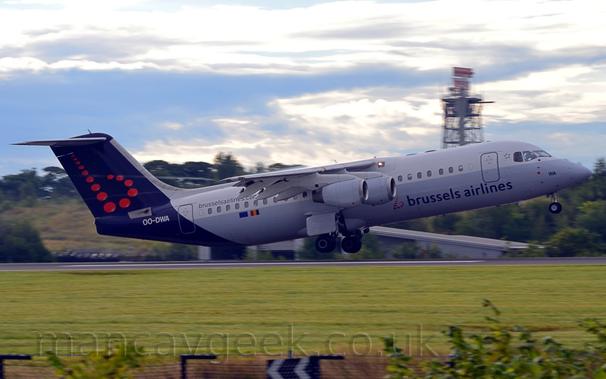 Side view of a high-winged, 4 engined jet airliner flying from left to right at a very low altitude, with undercarriage extended, flaps extended from the rear of the wings, and the nose raised sharply, suggesting it has just taken off.
The plane has a white upper half, with the lower half being grey, and black "Brussels Airlines" on the lower forward fuselage, ahead of a lower-case letter "b" made up of a series of red dots.
The black web address "BrusselsAirlines.com" is on the upper rear fuselage, over Belgian and EU flags.
The rear fuselage and tail are dark blue, with the white registration "OO-DWA" on the tail cone, and the lowercase "b" made of red dots on the tail.
Green grass fills the foreground, with trees on a small hill in the background, topped with a radar tower, under bright skies with grey and white clouds.