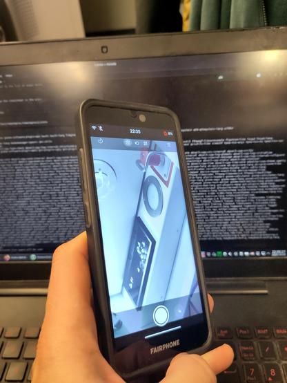 Fairphone 3, camera app preview with the front camera showing parts of a night train cabin. A laptop with the terminal open is visible behind the phone.