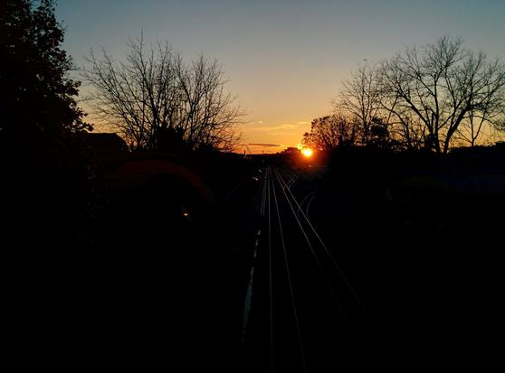 Short exposure (dark) photograph taken from a bridge overlooking the railway corridor in Kitchener, ON at sunset, looking west(ish) with the setting sun just north of the tracks. The foreground is unexposed, with only the tracks showing the reflection of the orange sunlight. Bare trees on either side of the track are sillhouetted against the sky. A few whispy clouds at the horizon accent the sky's gradient from a pale blue at the top of the frame to an orange glow at the horizon.