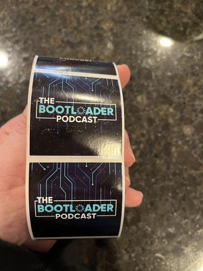 My hand holding a roll of stickers.  Two are face up that say "The Bootloader Podcast"