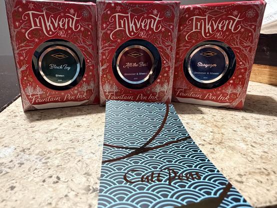 Three boxes of fountain pen ink in holiday-themed packaging are displayed. The inks are labeled as "Black Ivy," "All the best," and "Stargazer," each featuring descriptions related to sheen and shimmer. A blue decorative card with wave patterns and “cultpens” sits in the foreground