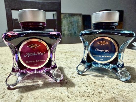 Two ink bottles are displayed on a countertop. The bottle on the left is labeled "All the Best" and features a purple hue, while the bottle on the right is labeled "Stargazer" and is blue. Both are from the brand Diamine