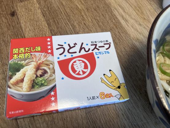 A packaged udon soup mix is displayed on a wooden surface. The box features a fox illustration, shrimp tempura, and text in Japanese. A bowl of udon with green onions is partially visible alongside the box.