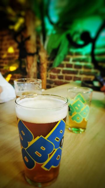 Un verre de bière re de Brussel Beer Project et deux autre derrière, sur une table de bois. En arrière plan un mur de brique avec un feuillage tropical.