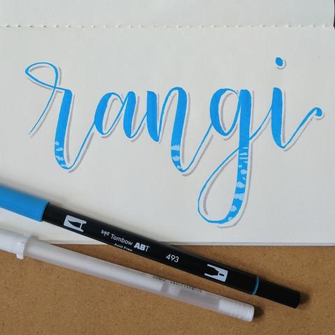on ivory paper is "rangi" (Maori for sky) written in bouncy, light blue calligraphy with a light grey shadow and some white puffy clouds in the lower parts of the letters.
below are a blue marker and white gel pen used to make this.